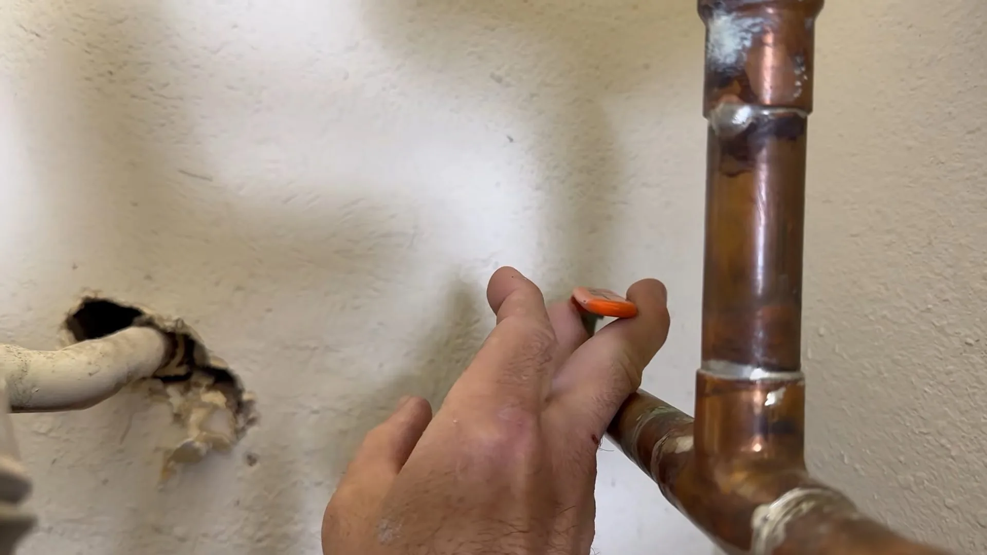 Hand holding a tool beside a soldered copper elbow at a hole cut in drywall