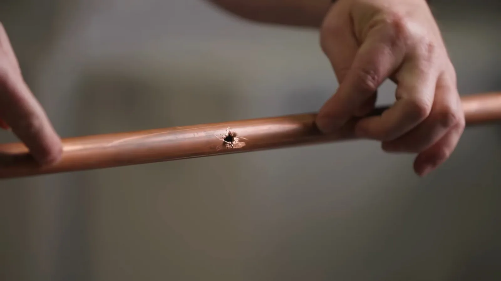 Two hands holding a length of copper pipe with a small pinhole in its wall