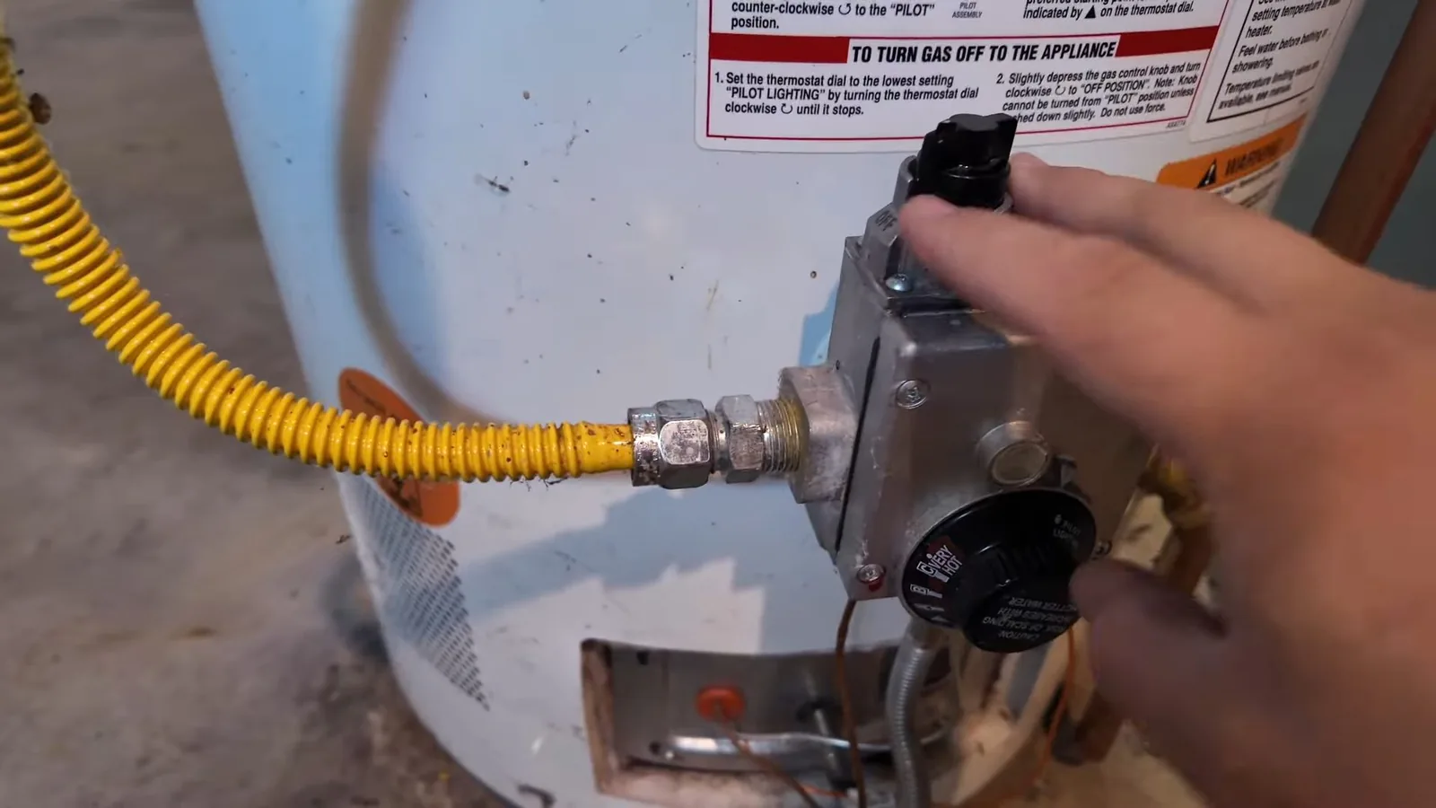 Hand on the gas control of a gas water heater, with a yellow flexible gas connector attached