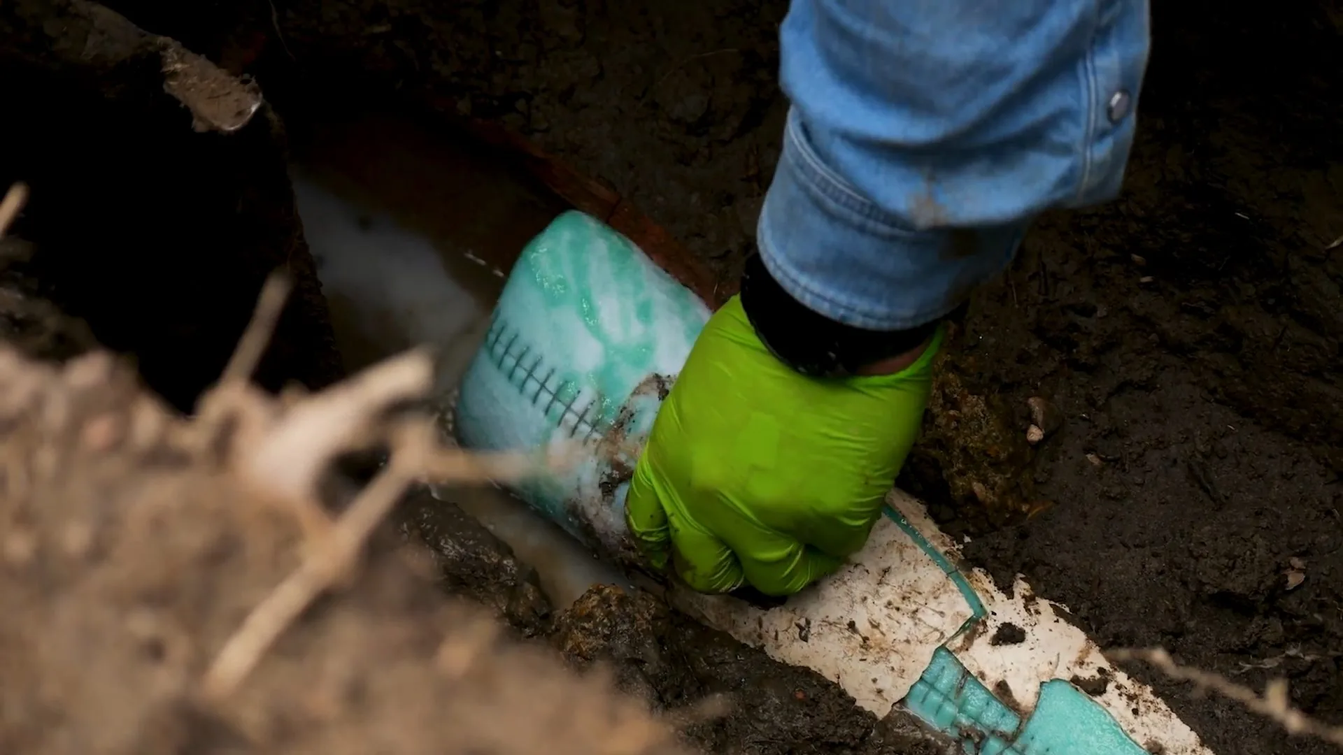 Green-gloved hand holding a white pipe section with a green patterned liner in a dug trench