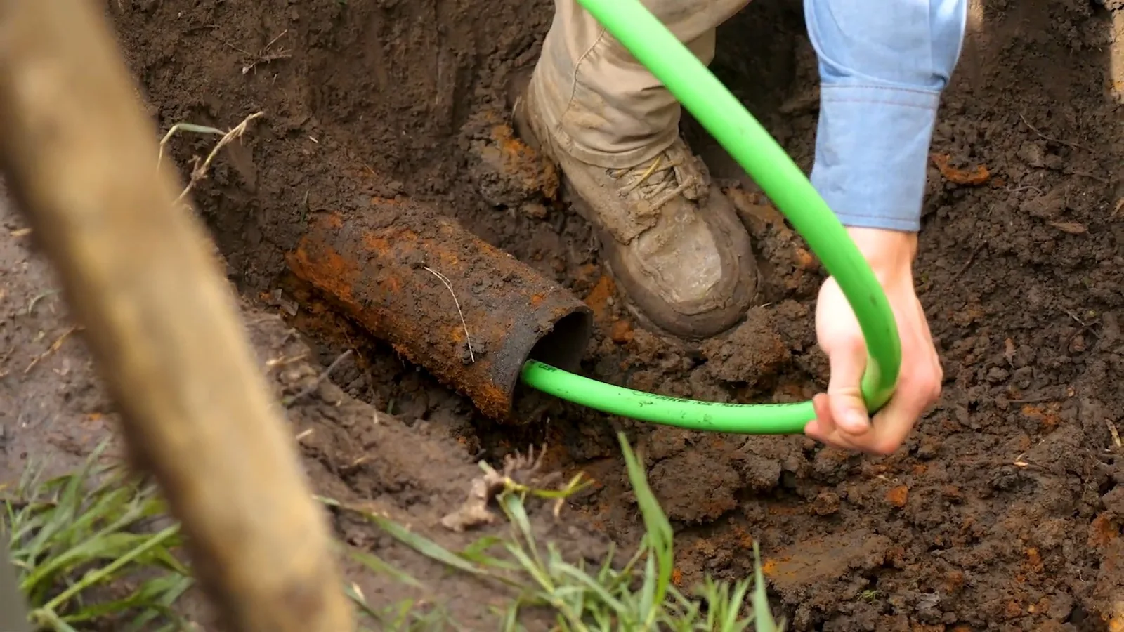 Green camera cable being fed into a rusty buried pipe in a dug trench