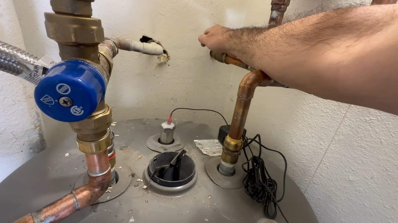 Hand at the top of a water heater beside new brass fittings, a blue valve and electrical wiring