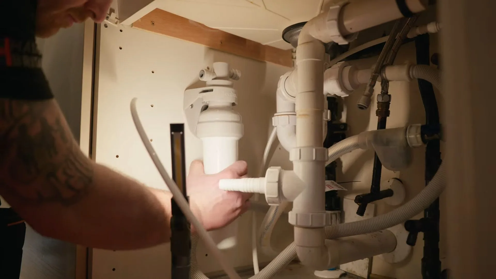 Hand holding the housing of a garbage disposal beside white drain pipes under a sink