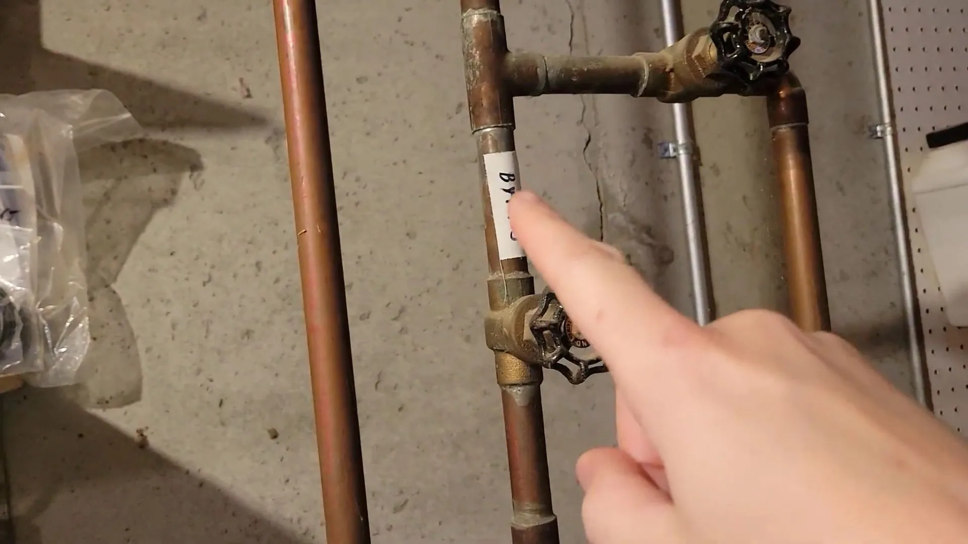 Hand pointing at two shutoff valves on copper supply pipes beside a basement wall