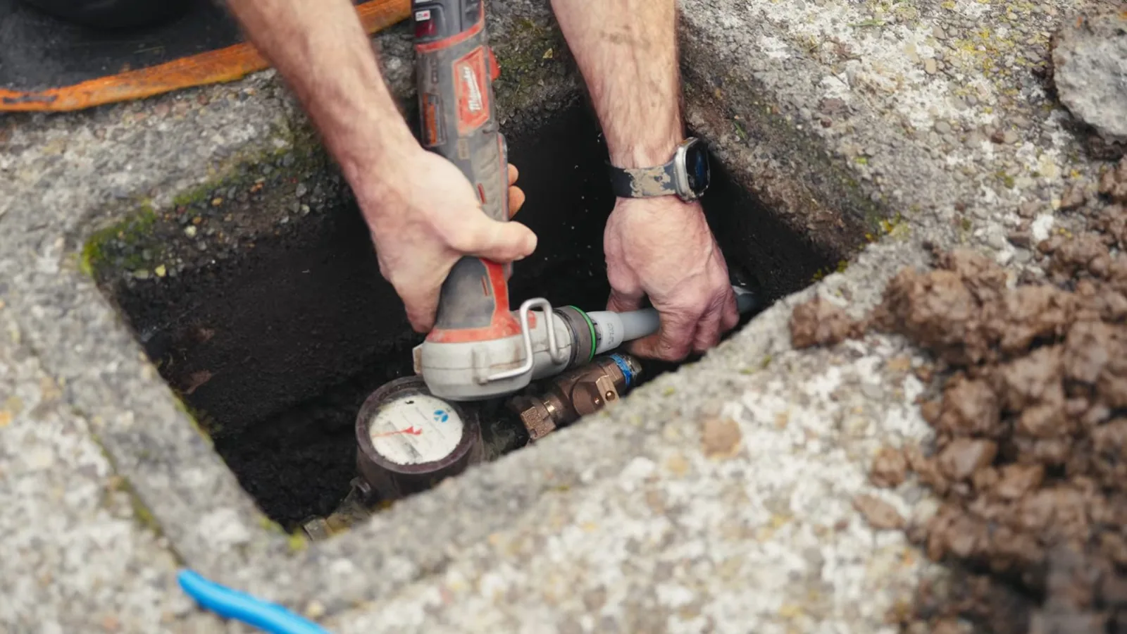 Hands working at a water meter inside an in-ground concrete box with a power tool