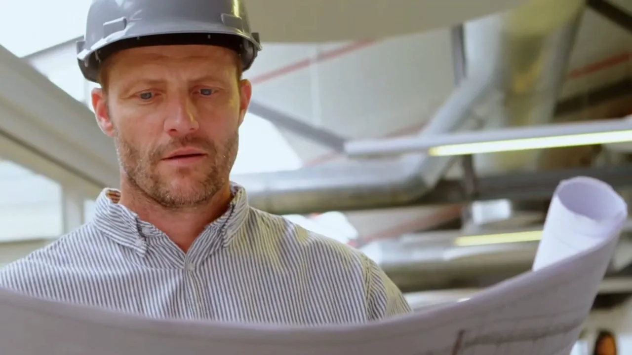 Man in a hard hat studying rolled plans under overhead ductwork