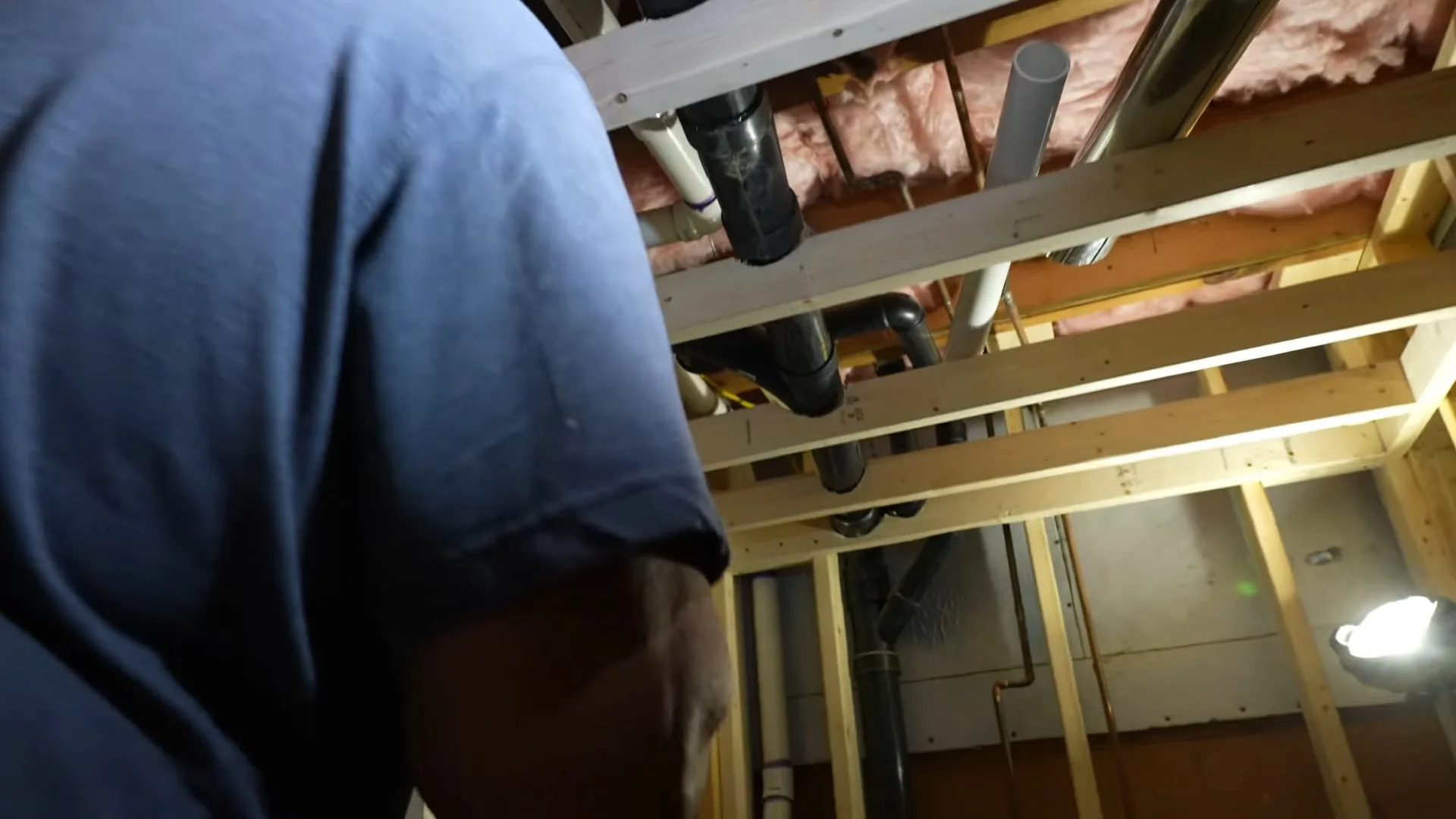 Man in a blue shirt looking up at drain and supply pipes between basement ceiling joists