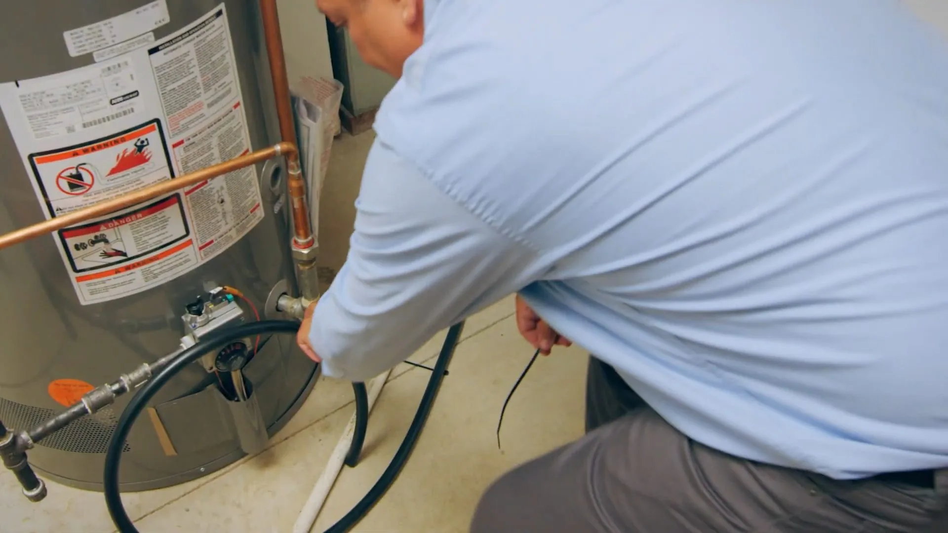 Man in a blue shirt crouched at a gas water heater working on its connectors
