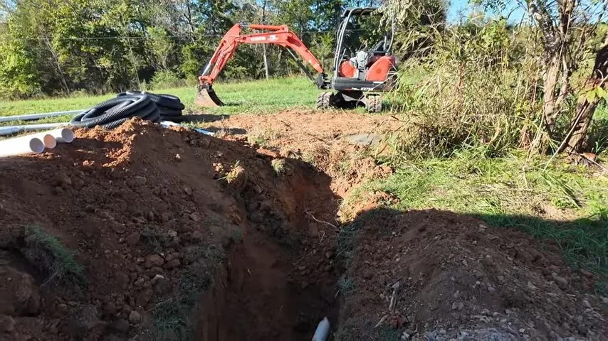 Mini excavator beside a long open trench with pipe laid on the grass