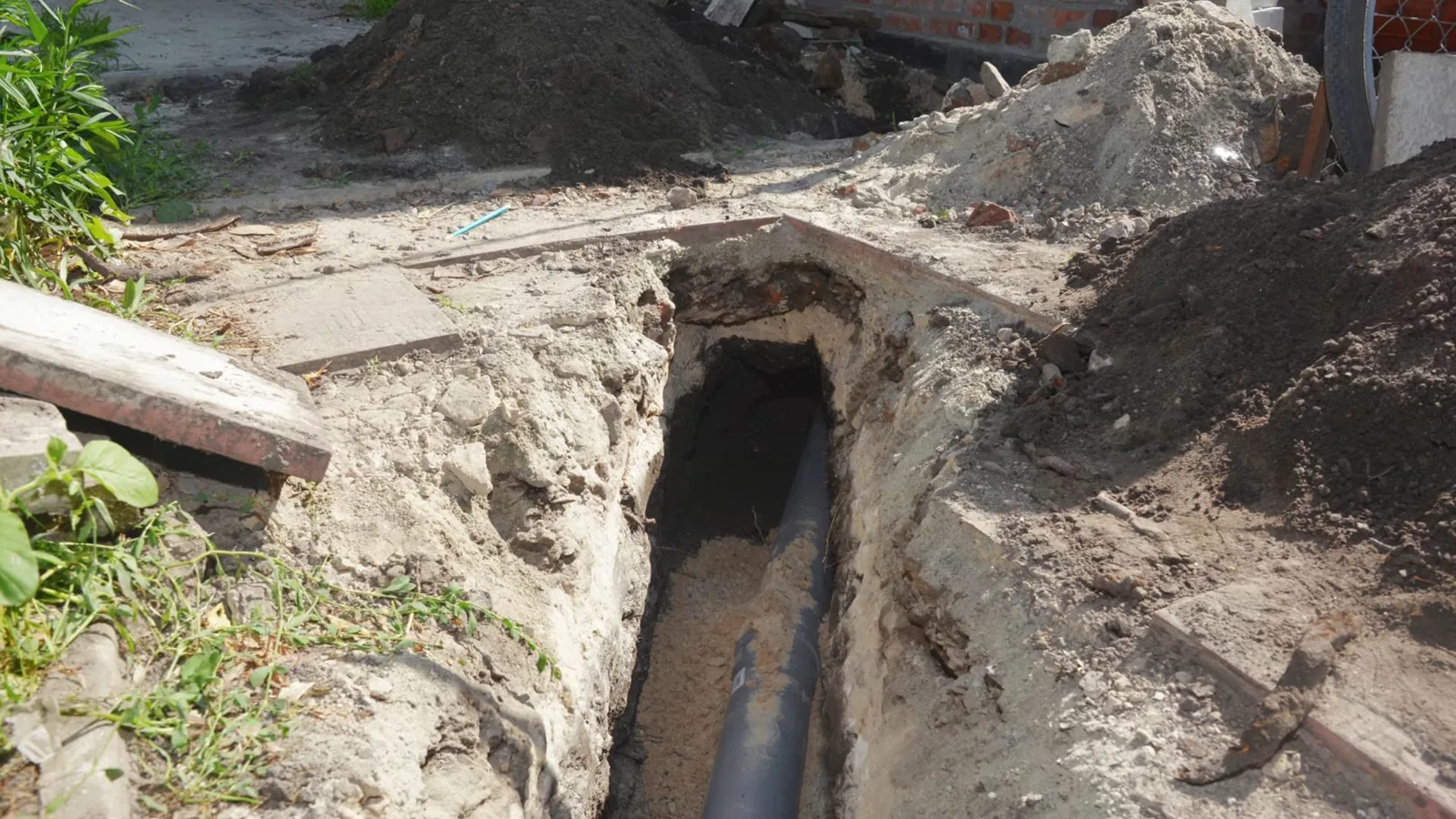 Narrow trench beside a house with a dark sewer pipe at the bottom and broken concrete at the edge