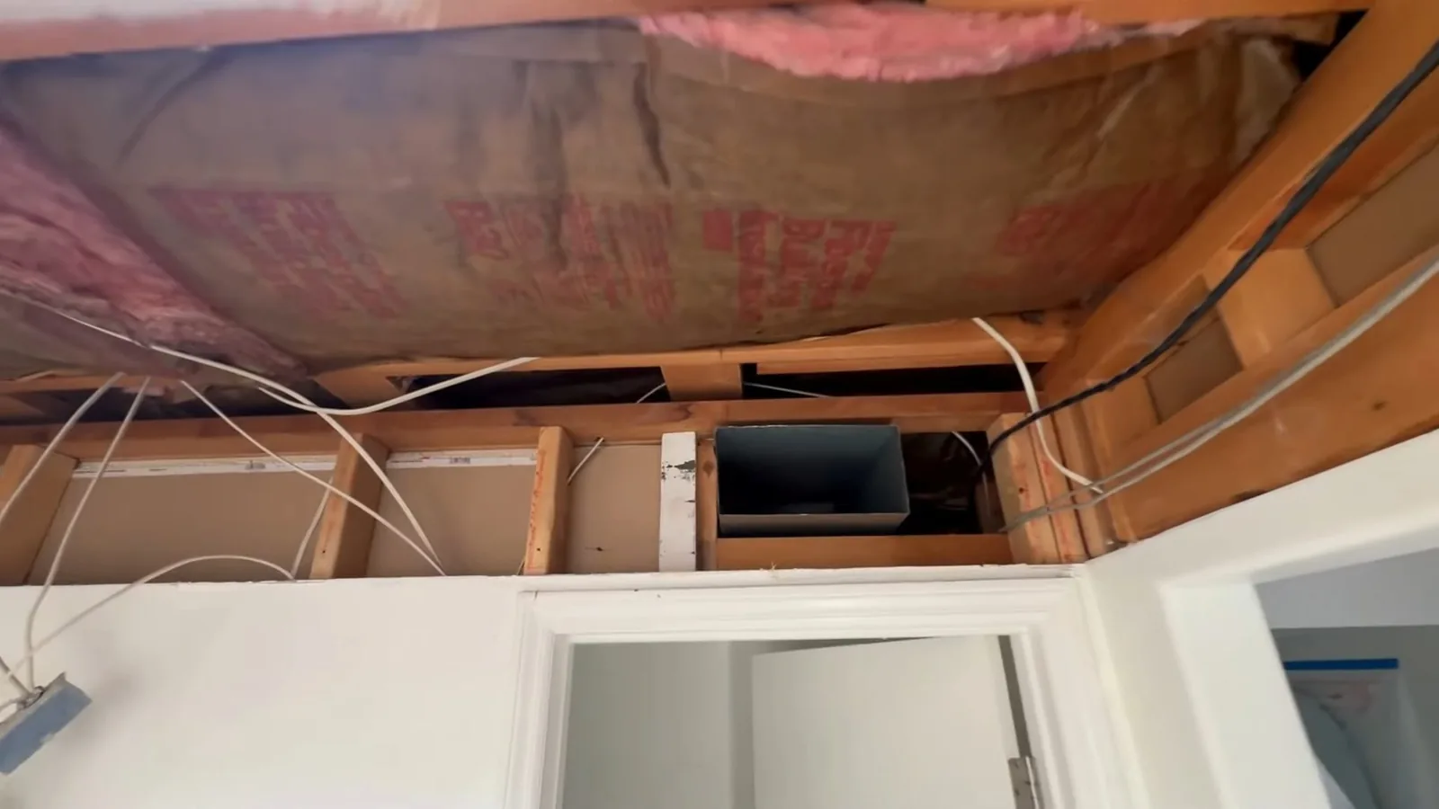 Opened ceiling showing pink insulation, joists and wiring above a door opening