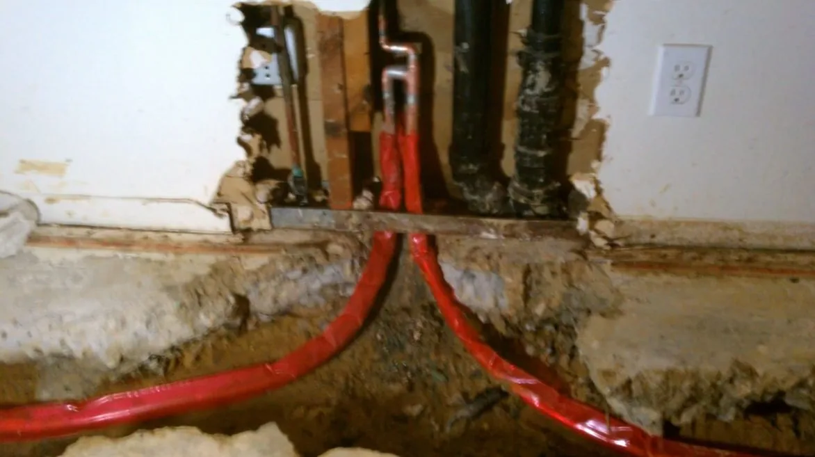 Opened floor edge showing red PEX tubing curving beside black cast iron pipe in the fill