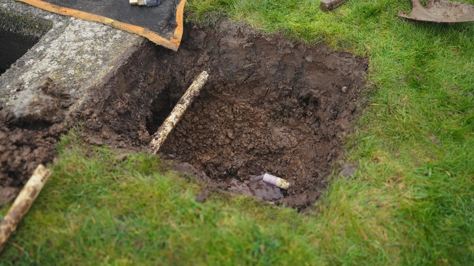 Square hole dug into a green lawn with a wooden stake lying in the soil