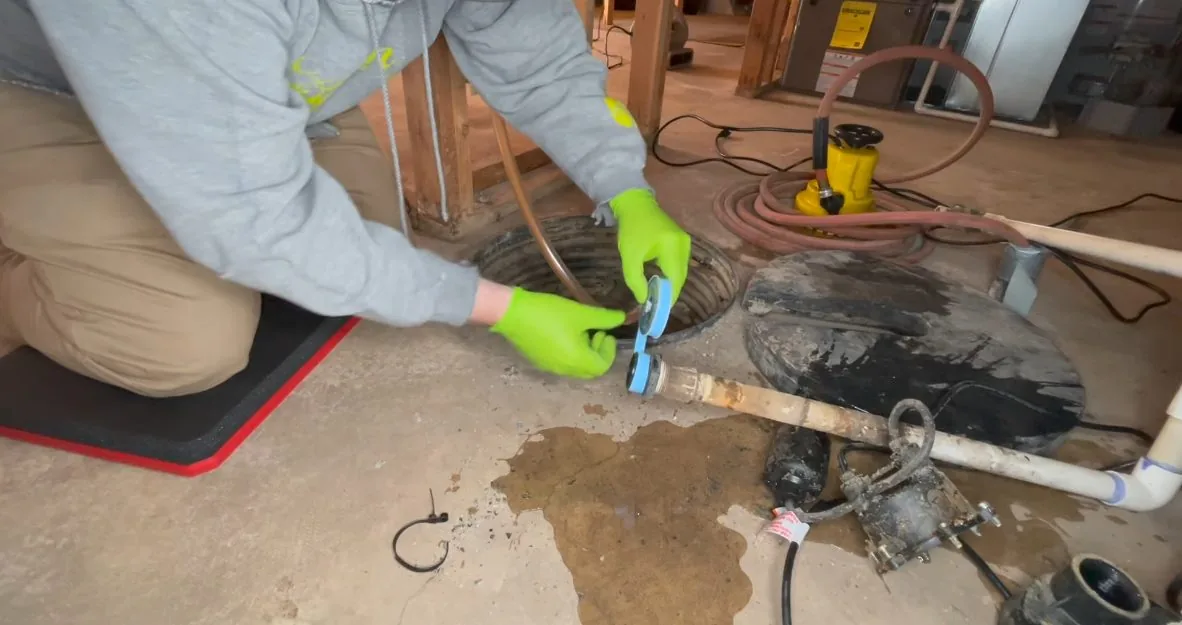 Man in green gloves kneeling at a floor drain beside a puddle on a concrete floor