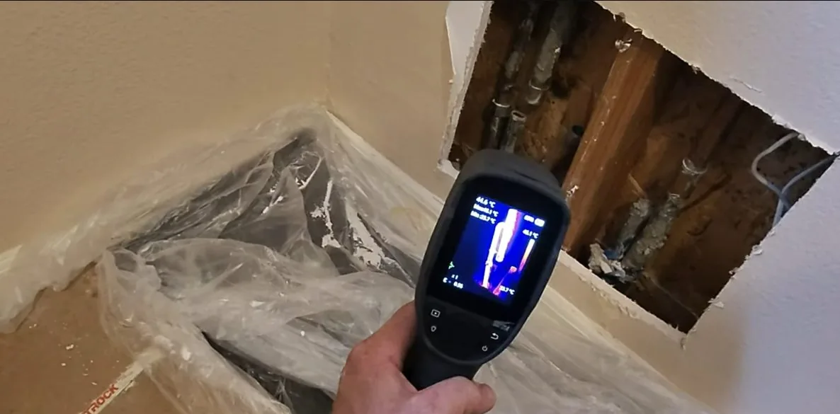 Thermal camera held toward an opened wall section showing pipes, with plastic sheeting on the floor
