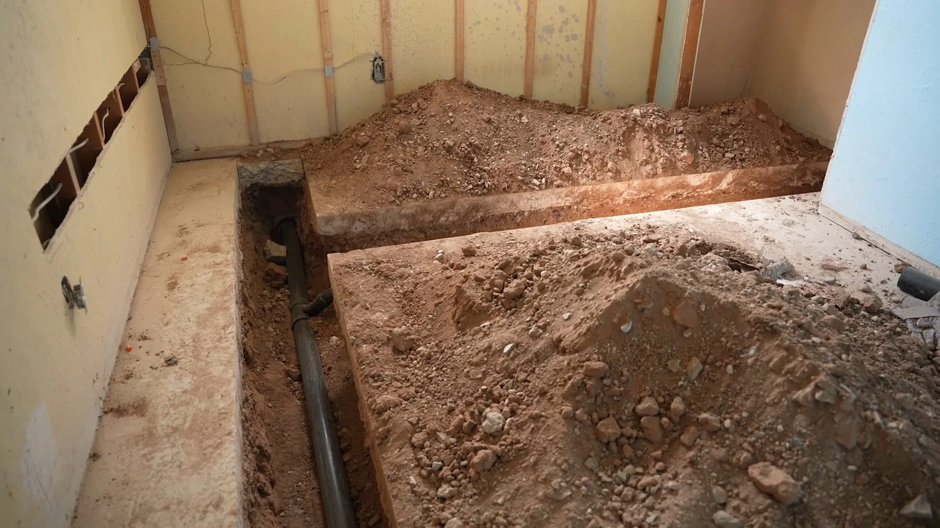 Interior room with a trench cut through the floor slab beside piles of excavated dirt and a dark pipe
