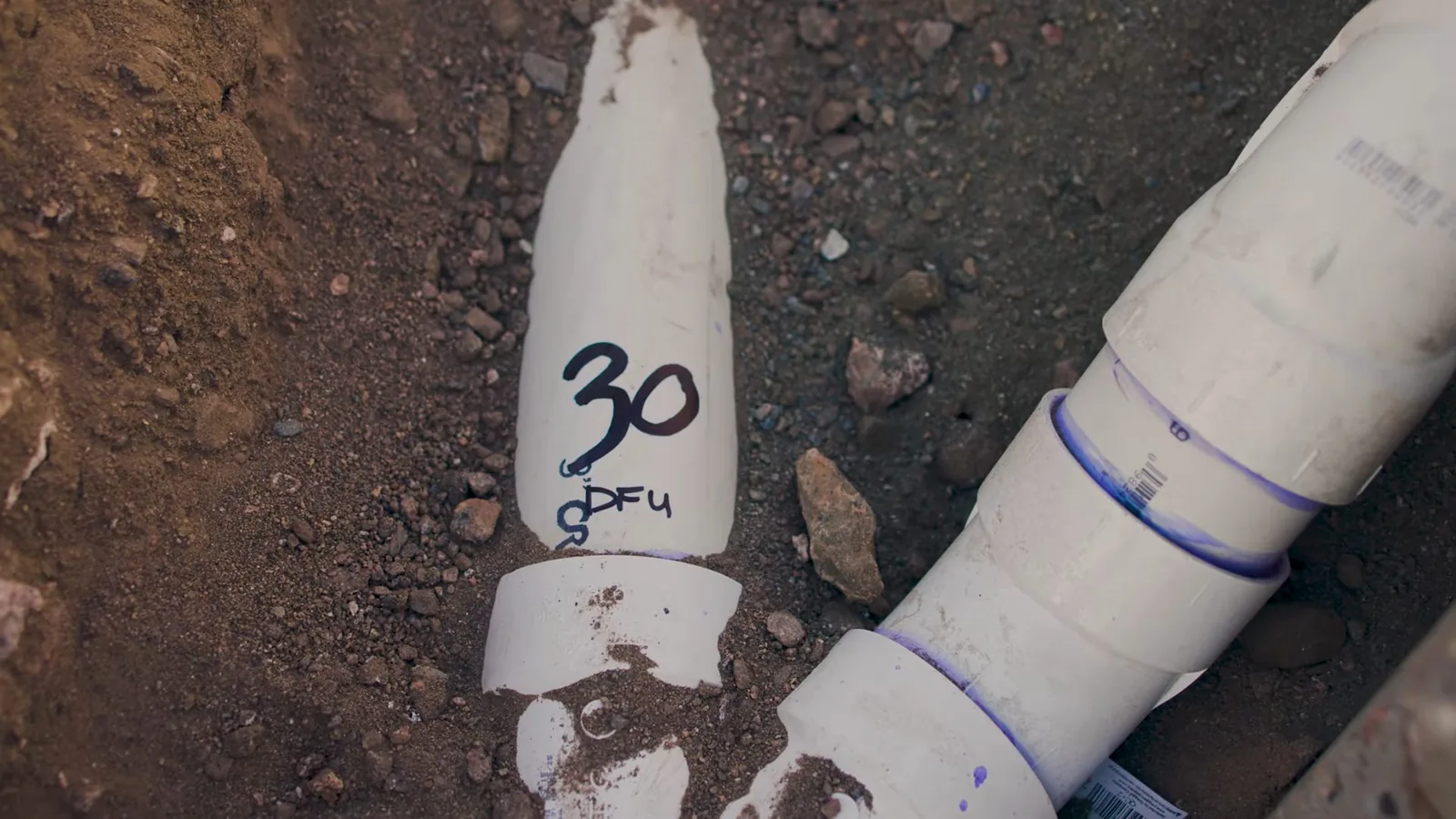 White PVC drain pipes with blue cement joints buried in soil, one marked with a number
