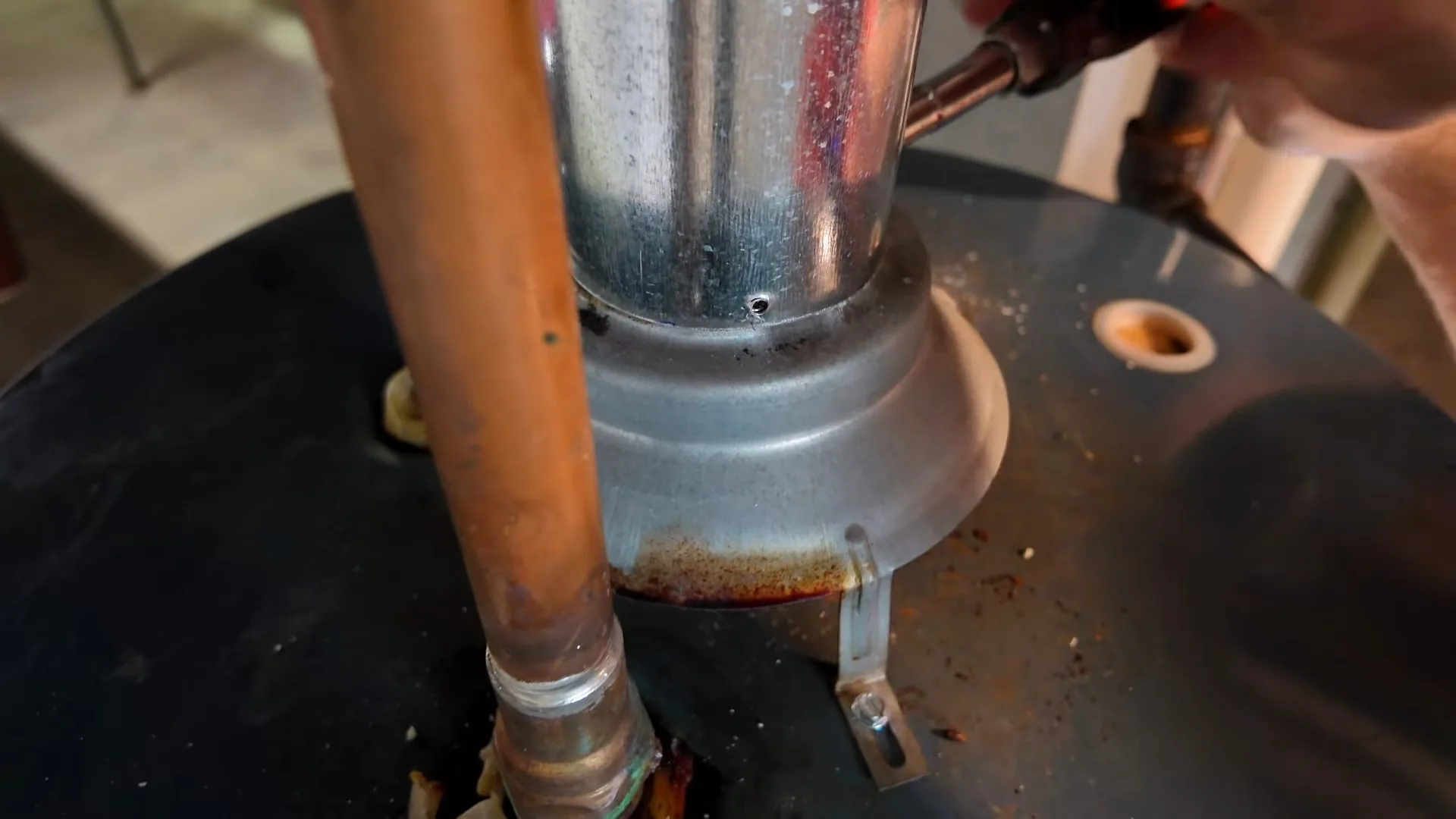 Close-up of a copper pipe and a metal flue collar on top of a water heater, with a technician's hand and torch at the right edge