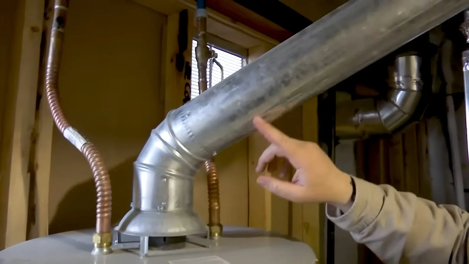 Hand pointing at the metal vent pipe and flexible connectors above a water heater in an unfinished basement