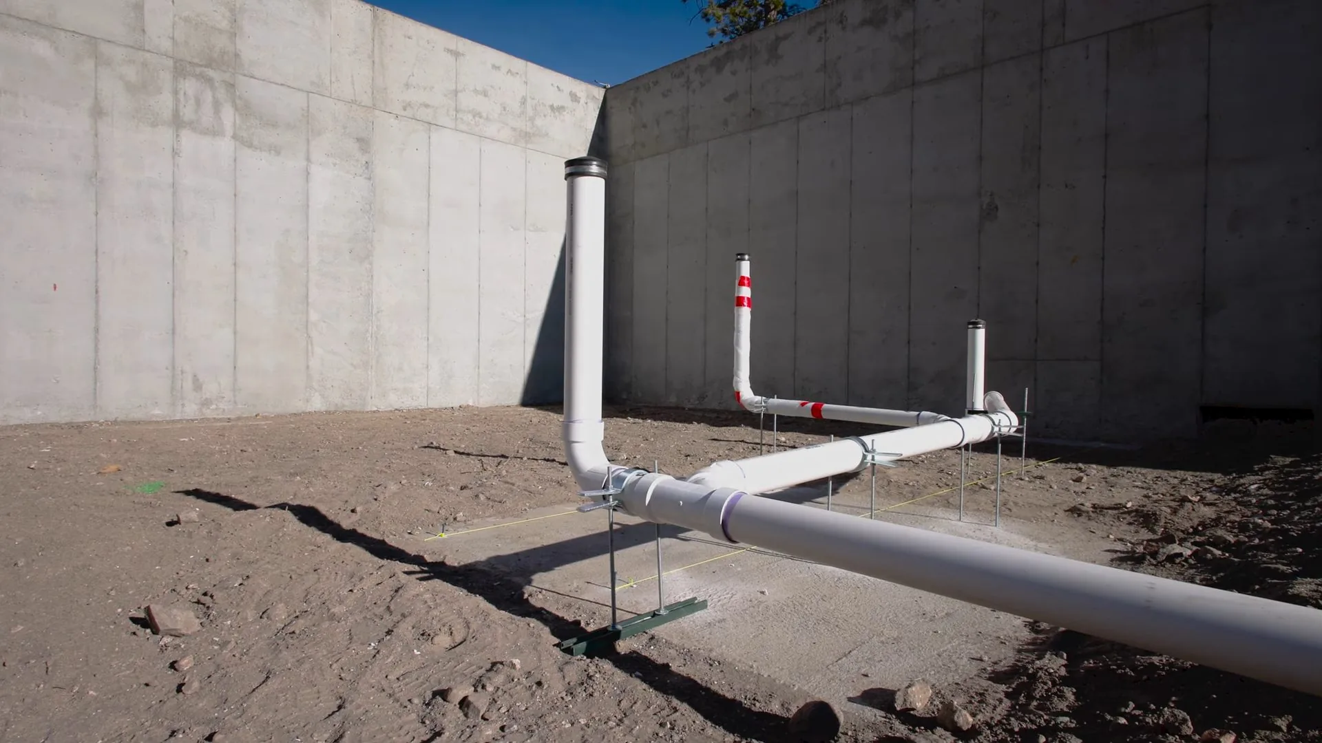 White drain pipes laid in bare soil and standing up beside a concrete wall