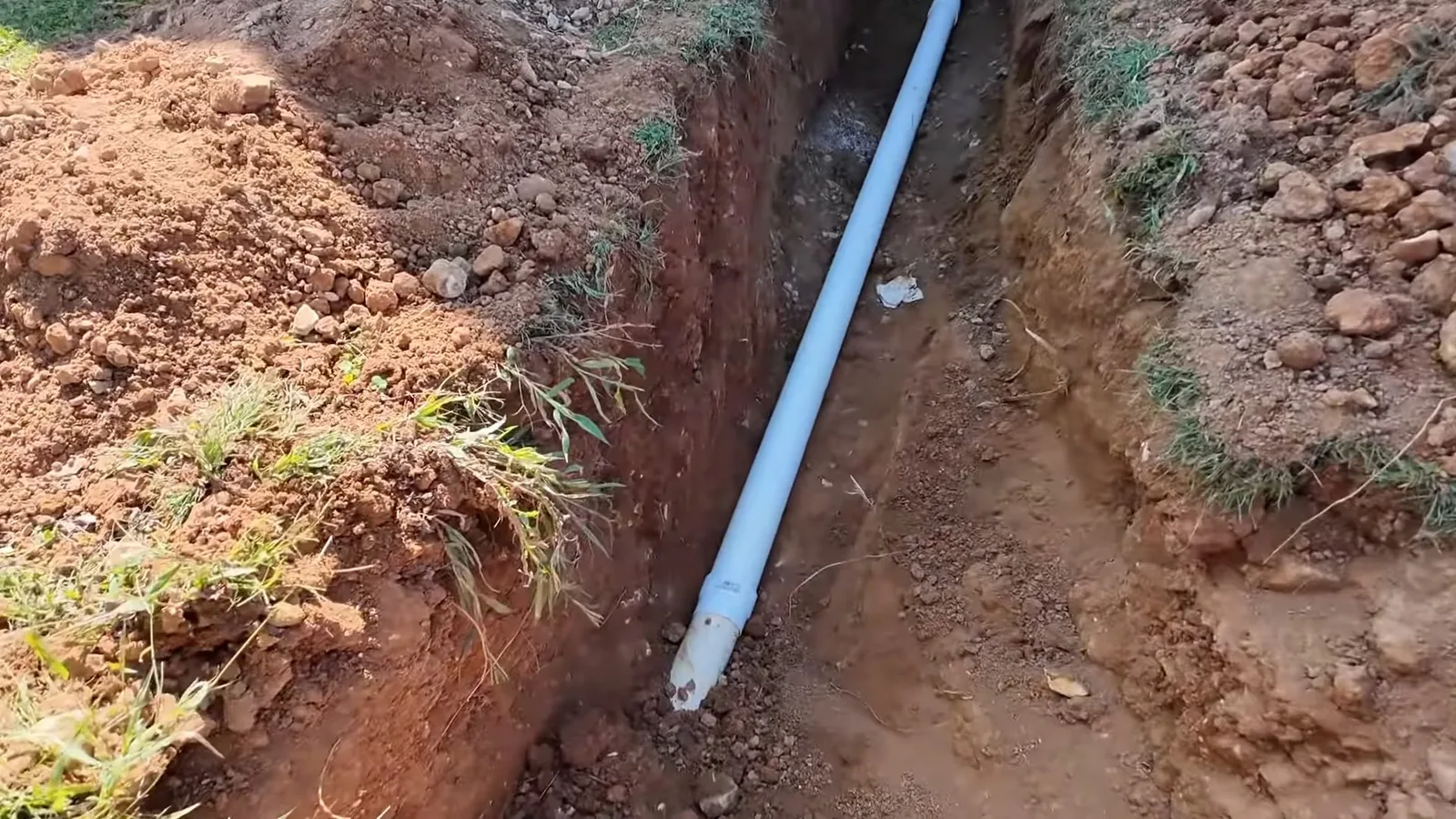 White pipe lying in a narrow dug trench in red clay soil