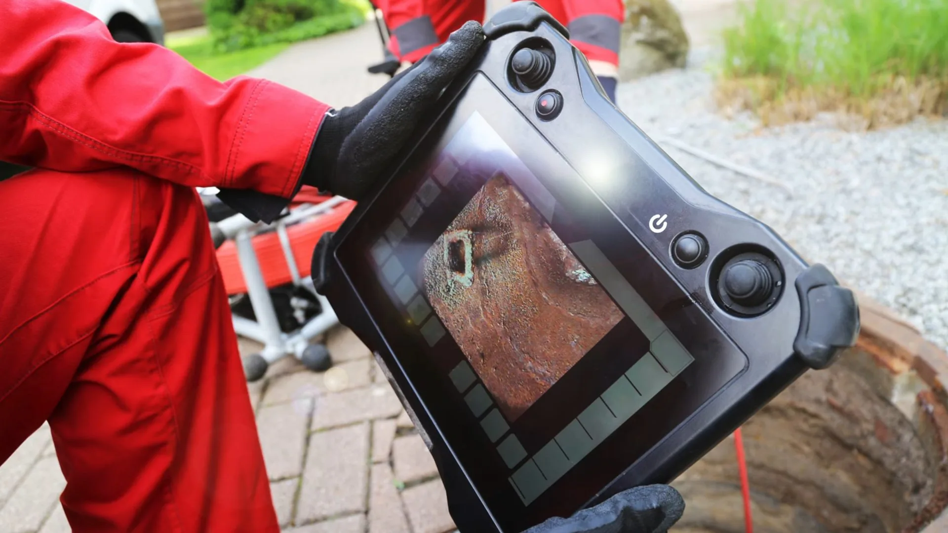 Person in a red coverall holding a camera monitor beside a paved patio
